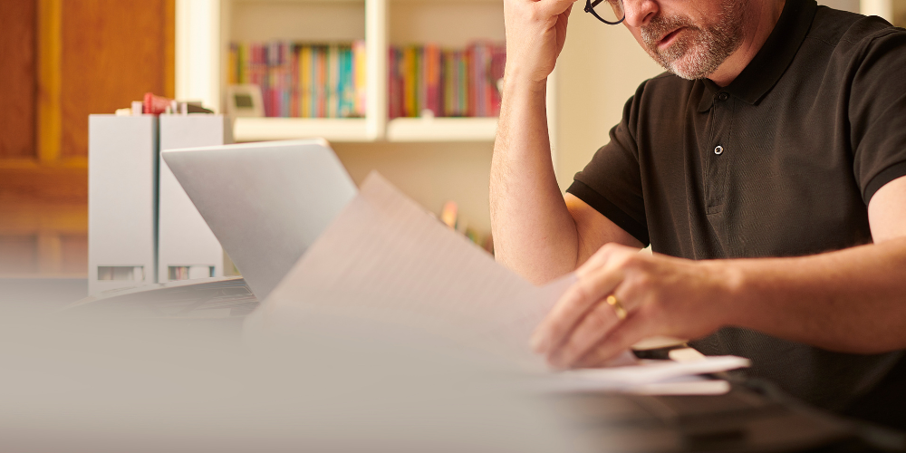 image of a man, sat at a table, with a laptop and papers in his hands. It is implied that the man is filing accounts for his small business